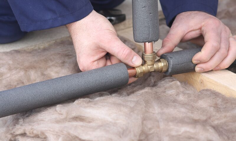 A person fitting grey foam insulation sleeves over copper pipes and fittings on loft insulation