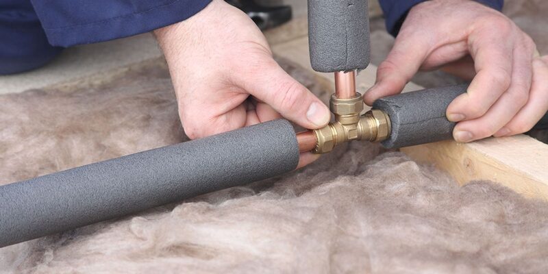 A person fitting grey foam insulation sleeves onto copper hot water pipes in a loft with mineral wool insulation