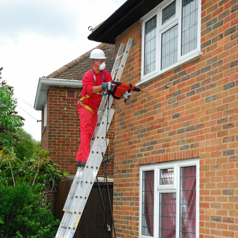 A worker in red overalls, hard hat and dust mask on a ladder drilling into the brick wall of a house with a red power drill, likely installing cavity wall insulation