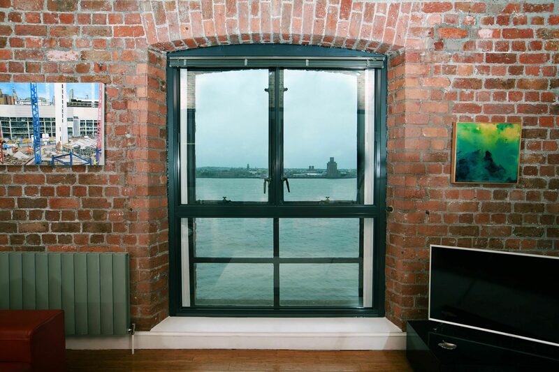 A hinged secondary glazing panel fitted inside an arched window in a brick-walled room overlooking water