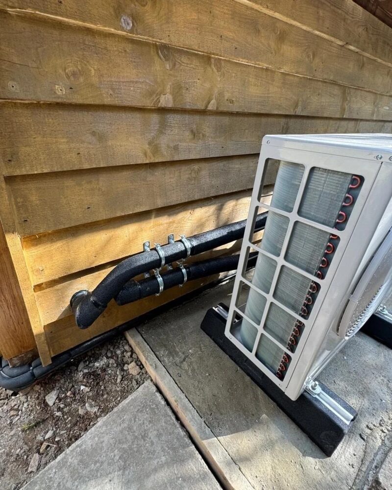 An outdoor air source heat pump unit with insulated refrigerant pipes entering a timber-clad exterior wall