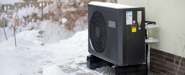 A black air source heat pump unit installed outside a house in snowy conditions, with insulated pipework running up the wall