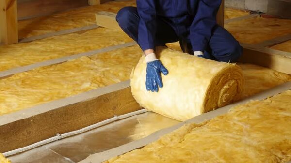 A worker in blue overalls and gloves unrolling a roll of yellow mineral wool loft insulation between wooden joists