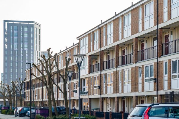 A row of brick social housing flats with balconies on a residential street, with a tall tower block behind and parked cars in front