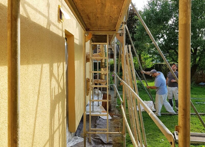 Workers erecting scaffolding against the yellow-rendered exterior wall of a house, apparently for external wall insulation or rendering work