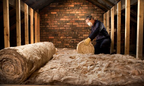 A worker wearing gloves and a dust mask unrolls mineral wool loft insulation between wooden joists in a brick-walled attic