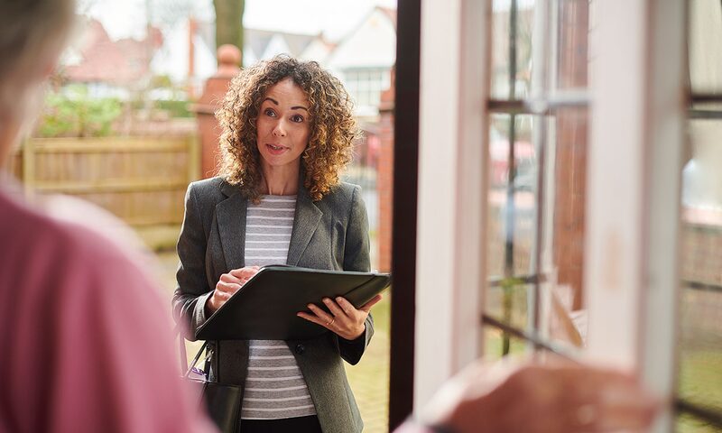 A woman holding a clipboard talking to a householder at their front door, as if cold calling