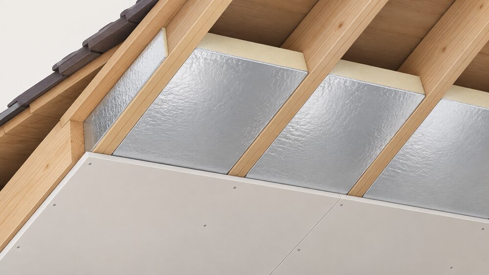 A close-up of a foil-faced rigid PIR insulation board fitted snugly between roof rafters, its shiny foil surface facing the viewer, with plasterboard being fitted beneath it and a gap-free, uncompressed fit visible at the joints.