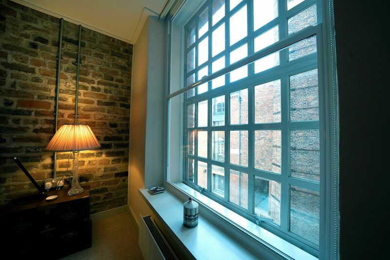 A secondary glazing panel installed inside a room beside a large multi-paned sash window, with a lamp on a cabinet against an exposed brick wall