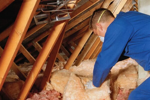 A worker in blue overalls and a dust mask laying mineral wool insulation between loft joists in an attic