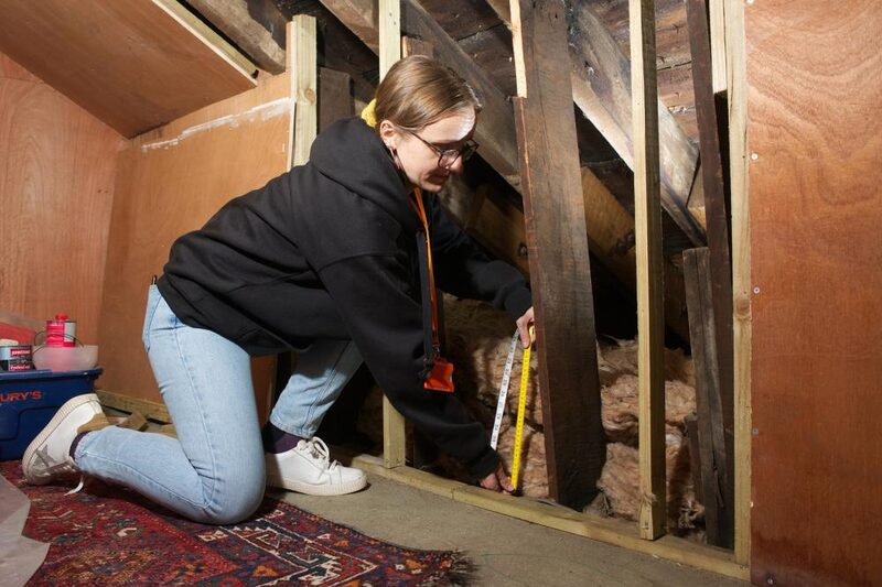 A woman kneeling in a loft measuring the depth of mineral wool insulation between wooden joists