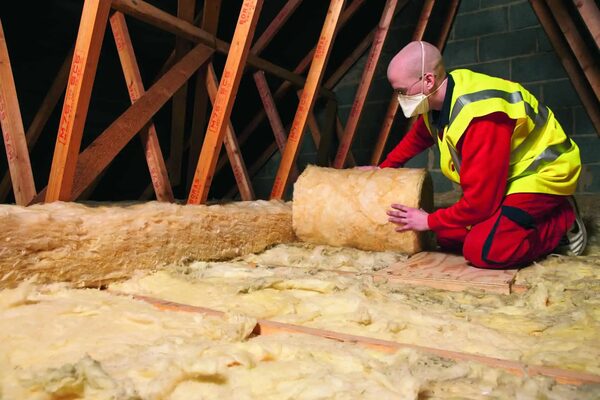 A worker in a hi-vis vest and dust mask unrolling a roll of mineral wool loft insulation between roof trusses in a loft