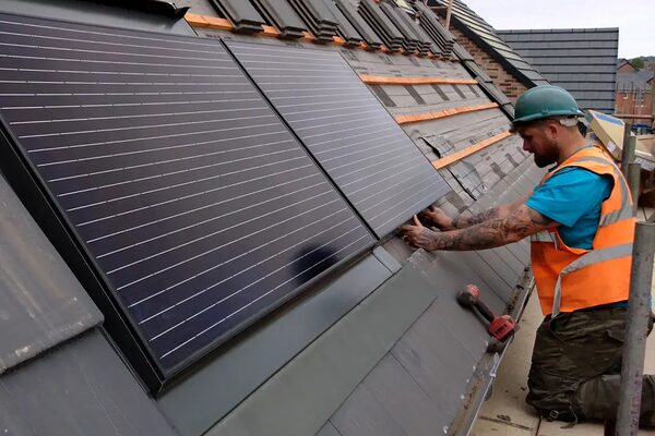 A roofer in a hard hat and hi-vis vest fitting solar panels onto a tiled roof