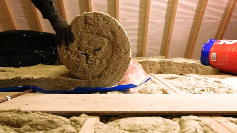 A gloved hand unrolling mineral wool loft insulation between joists in a loft, with a red and blue insulation roll wrapper at the side