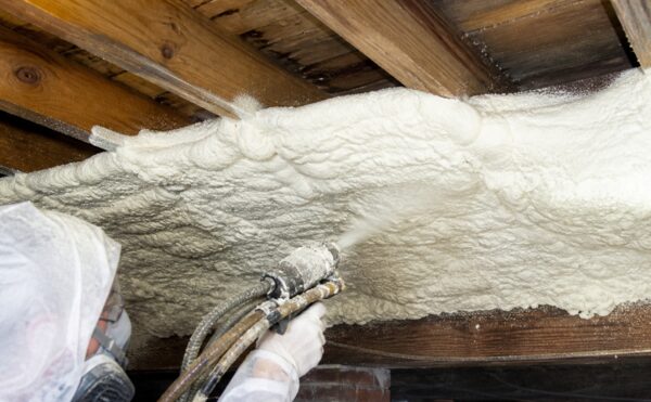 A worker in protective gear spraying white spray foam insulation between wooden floor joists