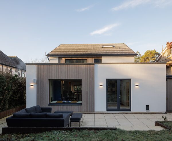 A modern Passivhaus retrofit house extension with timber cladding, white render, large glazing and a paved patio with outdoor sofa