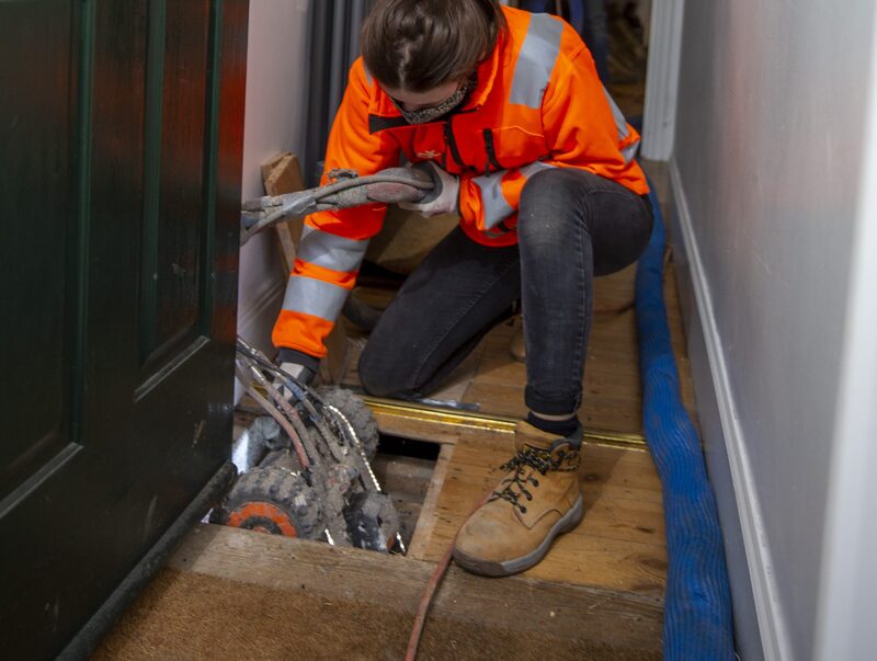 A Q-Bot installer in a hi-vis orange jacket lowering an insulation robot through a floorboard hatch in a home hallway