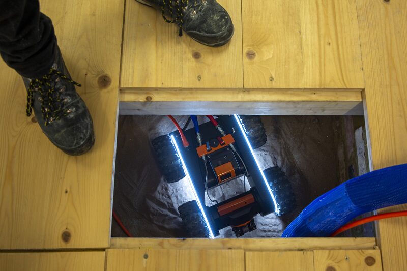 A Q-Bot underfloor insulation robot being lowered through a hatch in a suspended timber floor, with a worker's boots beside the opening