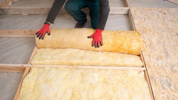 A worker in red gloves unrolling yellow mineral wool insulation between wooden floor joists