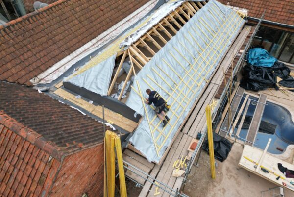 A roofer installing silver multifoil insulation over the rafters of a partially stripped pitched roof on a house under renovation