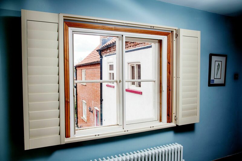 A horizontal sliding secondary glazing panel installed inside a sash window with cream shutters and a radiator below, in a blue-walled room