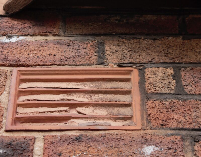 A terracotta air brick vent partially blocked with debris, set into a red brick wall