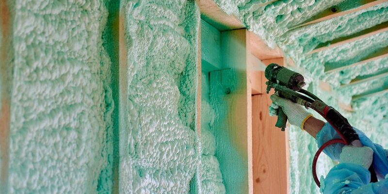 A worker in gloves spraying green spray foam insulation onto the wooden rafters and walls of a loft under construction