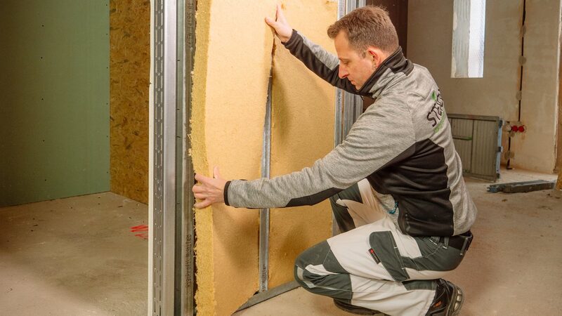 A tradesman fitting a wood-fibre internal wall insulation board into a metal stud frame during a renovation