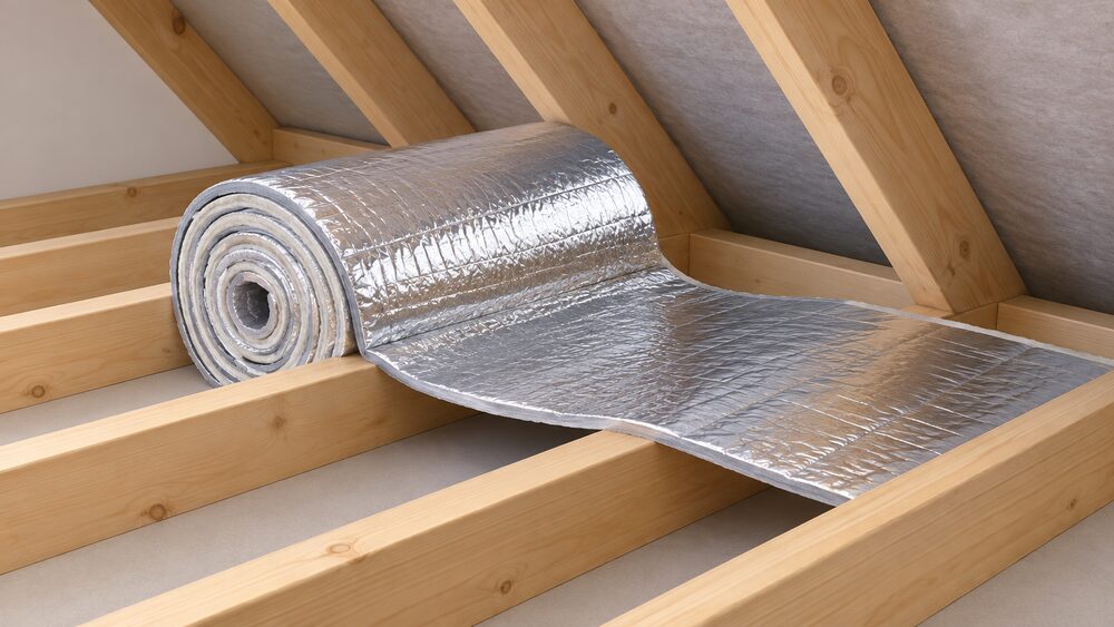 A close-up of a partially unrolled roll of reflective multifoil insulation, its cut end showing the layered foil faces and wadding, draped between loft joists under a pitched roof with a visible air gap left in front of the foil surface.