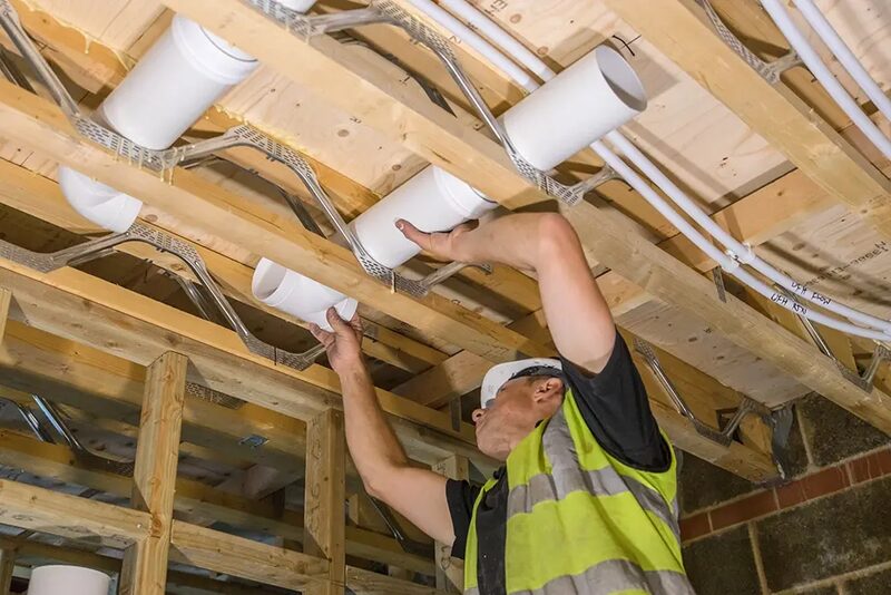 A worker in a hi-vis vest and hard hat installing white cylindrical ventilation ducting between timber floor joists