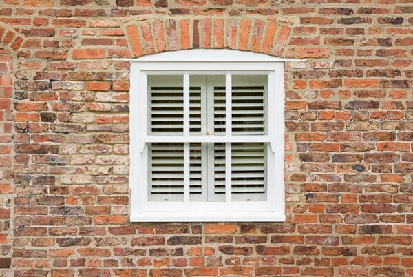 A white sash window with shutters set in a red brick wall with an arched brick lintel