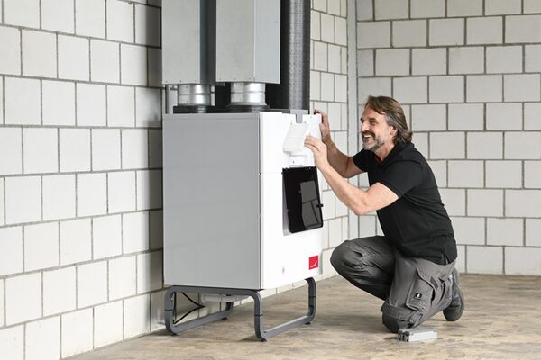 A technician kneeling beside a white Zehnder heat recovery ventilation unit installed against a white block wall