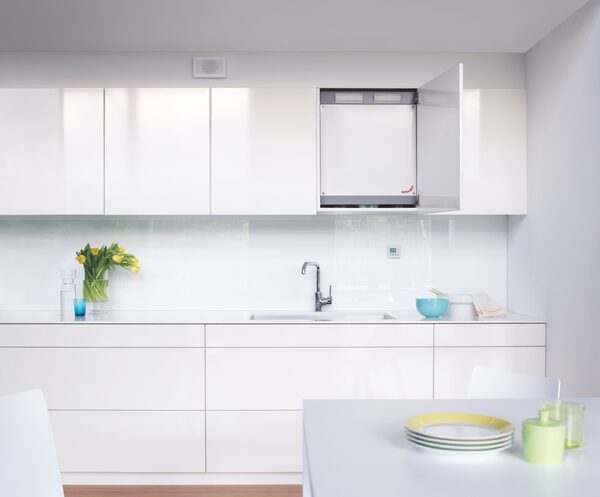 A Zehnder ComfoAir 180 mechanical ventilation unit installed inside a white kitchen wall cupboard above the worktop
