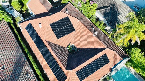 Aerial view of a terracotta-tiled house roof fitted with several arrays of black solar panels, with a swimming pool and neighbouring homes nearby