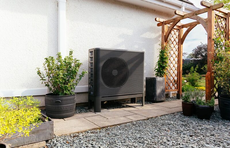 An Aira air source heat pump unit installed outside a white-walled house beside a garden path and wooden pergola