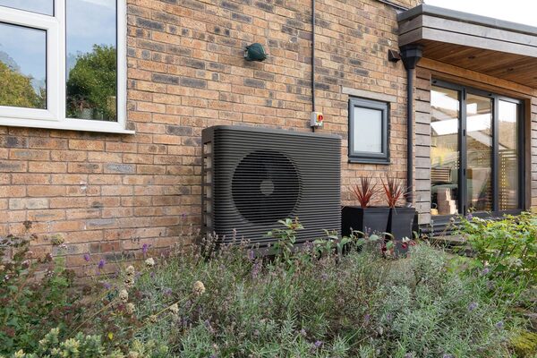 An Aira air source heat pump installed on the brick exterior wall of a home beside a garden