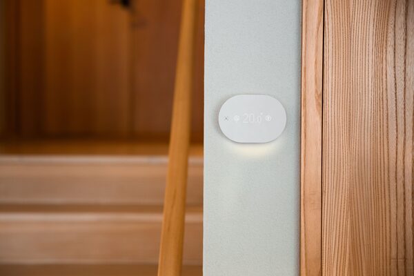 A white oval Aira smart thermostat mounted on a pale wall showing 20.0 degrees, next to wooden stairs