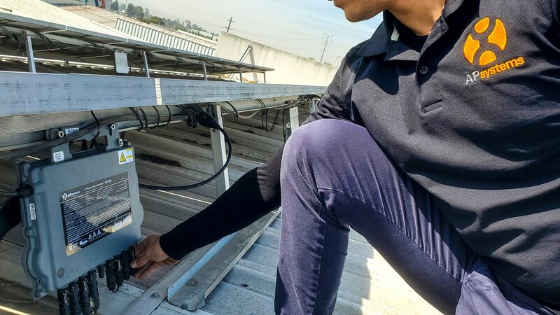 A technician in an APsystems polo shirt installing an APsystems microinverter beneath solar panels on a rooftop
