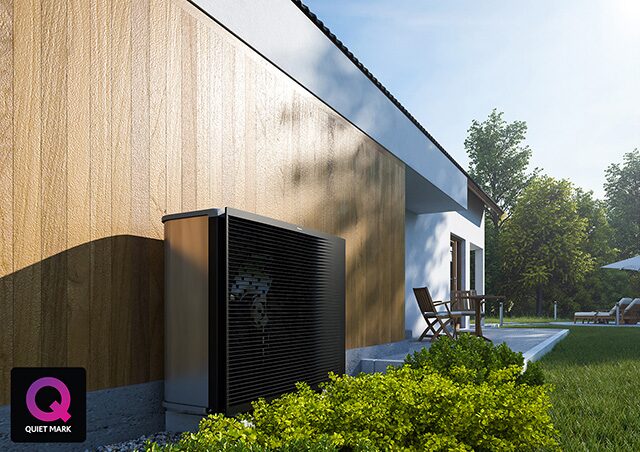 A dark air source heat pump unit installed on the outside wall of a modern timber-clad house beside a patio