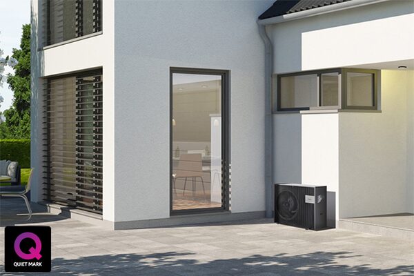 A black air source heat pump unit installed outside a modern white house on a paved patio, with a Quiet Mark logo in the corner