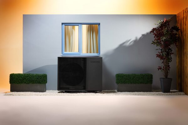 A Samsung air source heat pump unit installed on a wall below a window in a staged outdoor setting with planters
