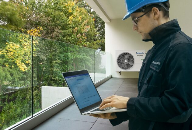 A Panasonic engineer in a hard hat using a laptop next to a wall-mounted Panasonic air source heat pump on a balcony