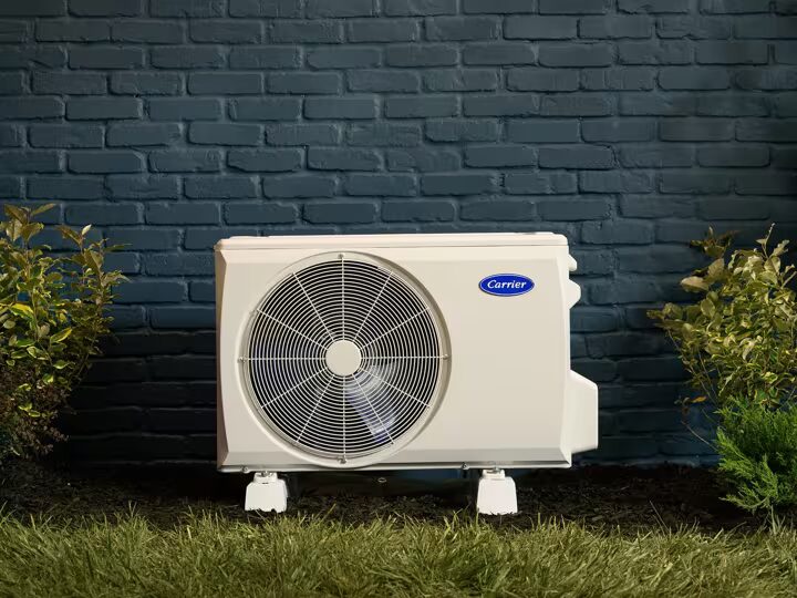 A Carrier outdoor heat pump unit installed on feet against a dark blue brick wall beside shrubs
