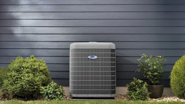 A Carrier outdoor heat pump unit installed against a dark blue-sided house wall with shrubs around it