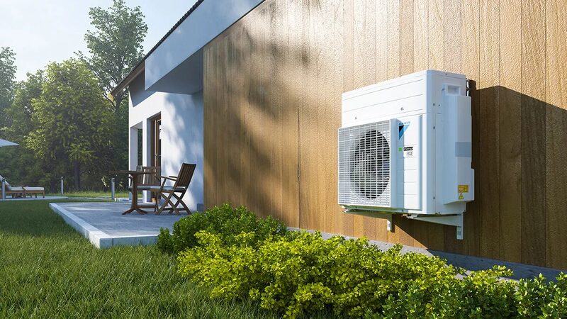 A Daikin heat pump outdoor unit mounted on the wooden exterior wall of a house beside a garden patio
