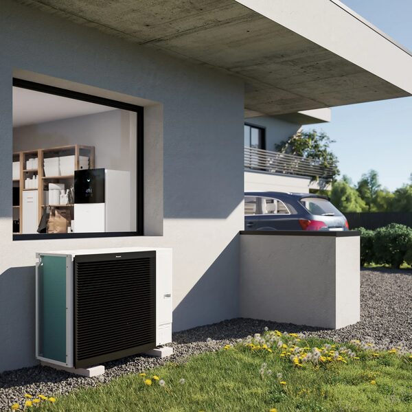 A Daikin air source heat pump outdoor unit installed beside a modern house, with an indoor unit visible through the window