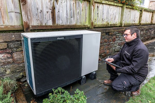 An engineer in a Daikin jacket inspects an installed Daikin air-to-water heat pump unit in a garden beside a stone wall