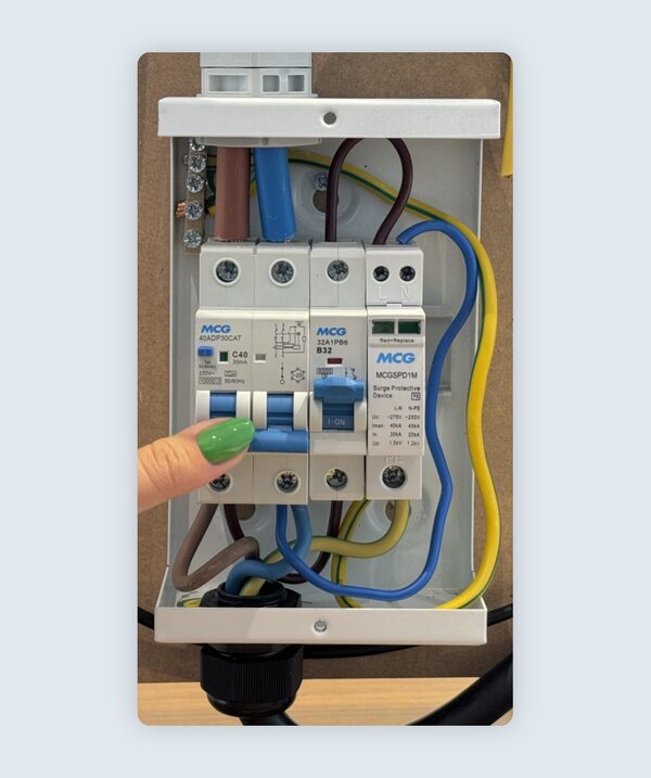 A finger pressing the reset switch on an MCG RCBO and surge protection device inside a consumer unit with exposed wiring