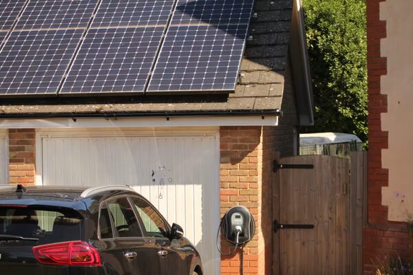 A brick house with rooftop solar panels and an EV charge point on the wall charging a dark car outside a garage