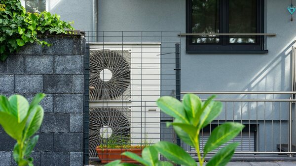 An air source heat pump unit with two fans installed outside a grey house behind a metal guard fence